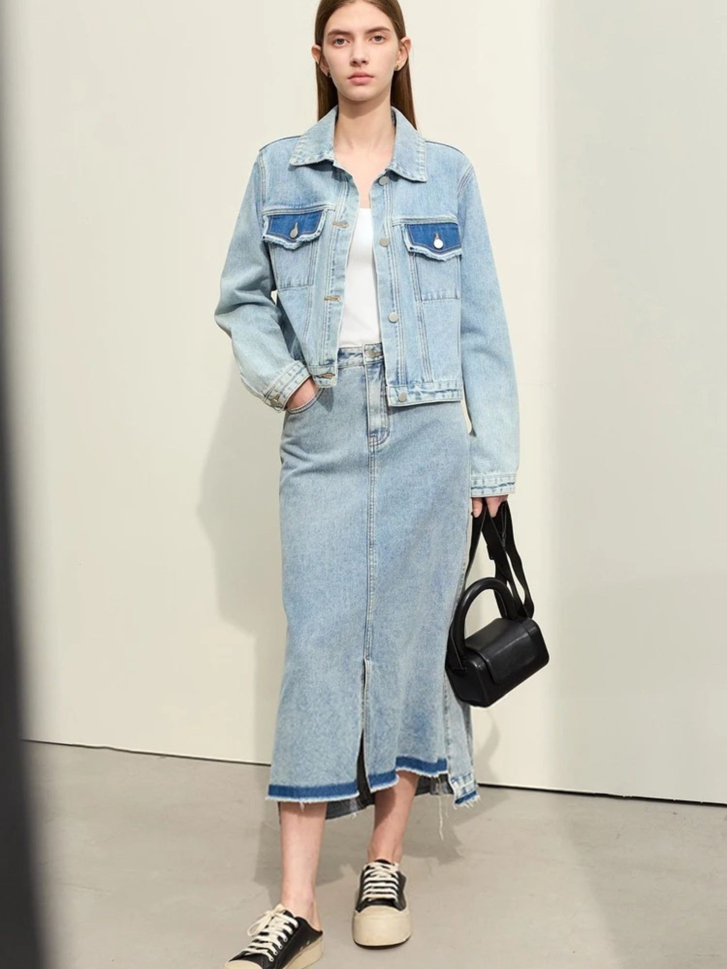 Woman wearing a denim jacket and skirt with a black handbag on a plain background