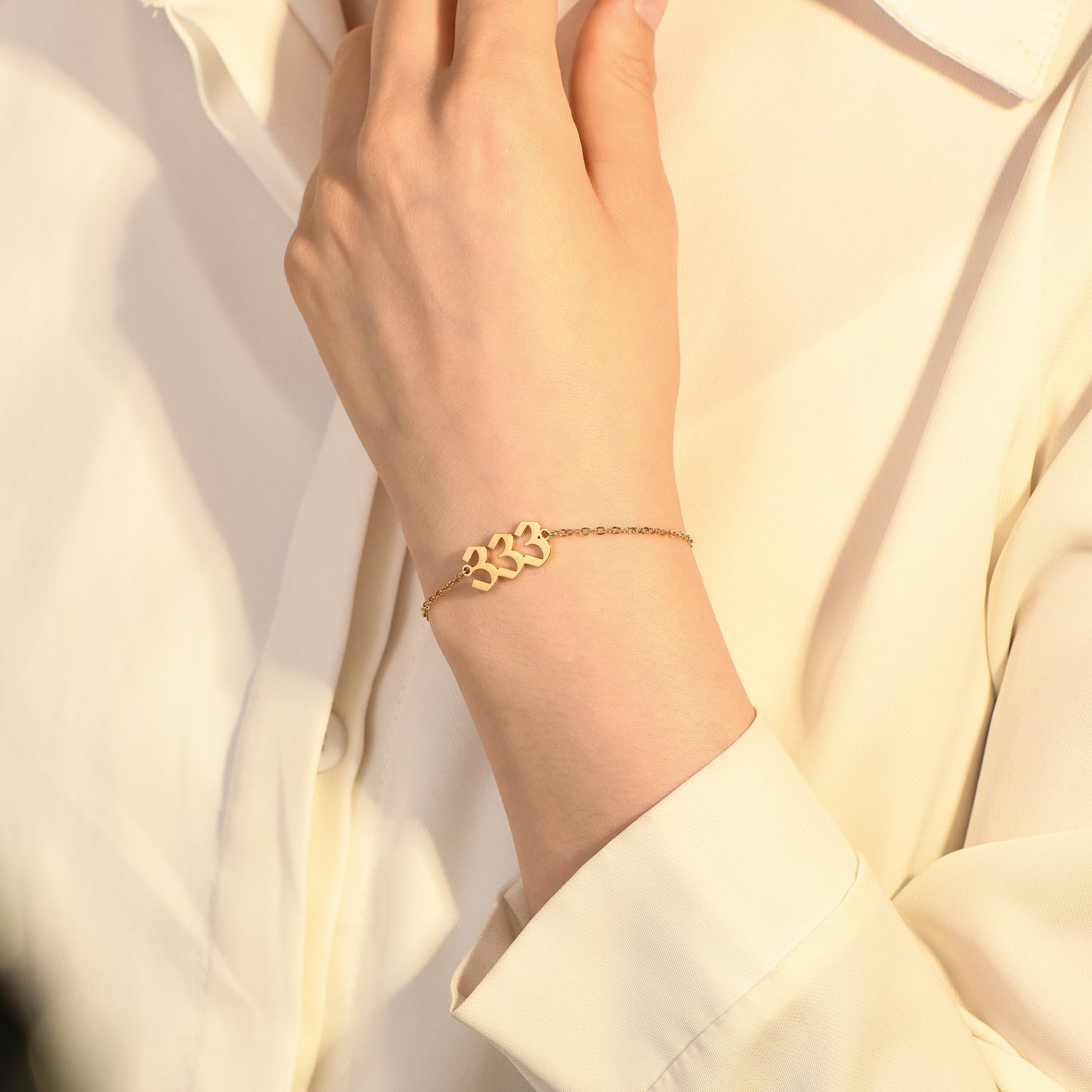 Gold angel number 333 bracelet on woman's wrist wearing a white long sleeve top