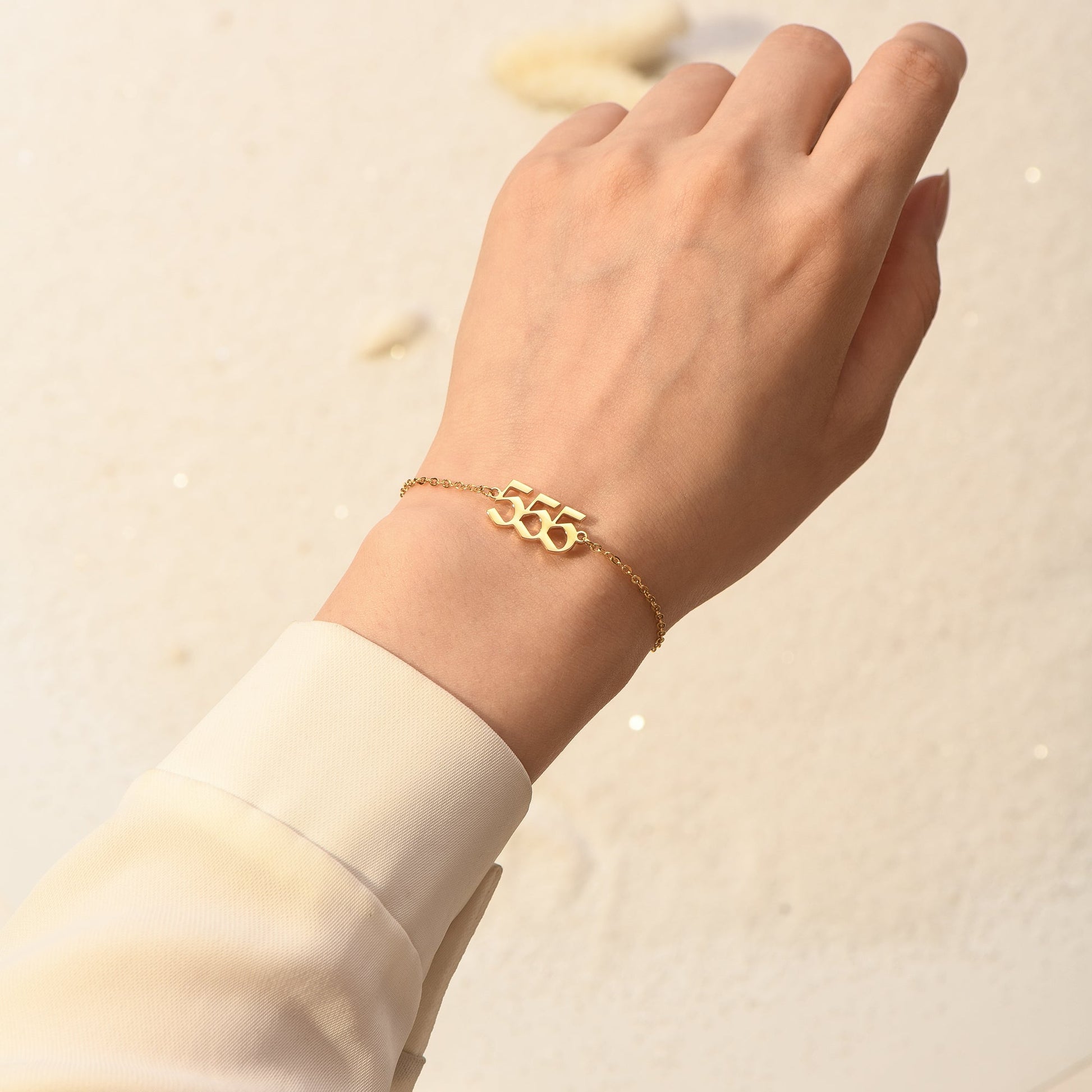 Gold bracelet with angel number 555 on a woman's wrist who's wearing white long sleeve with a white background