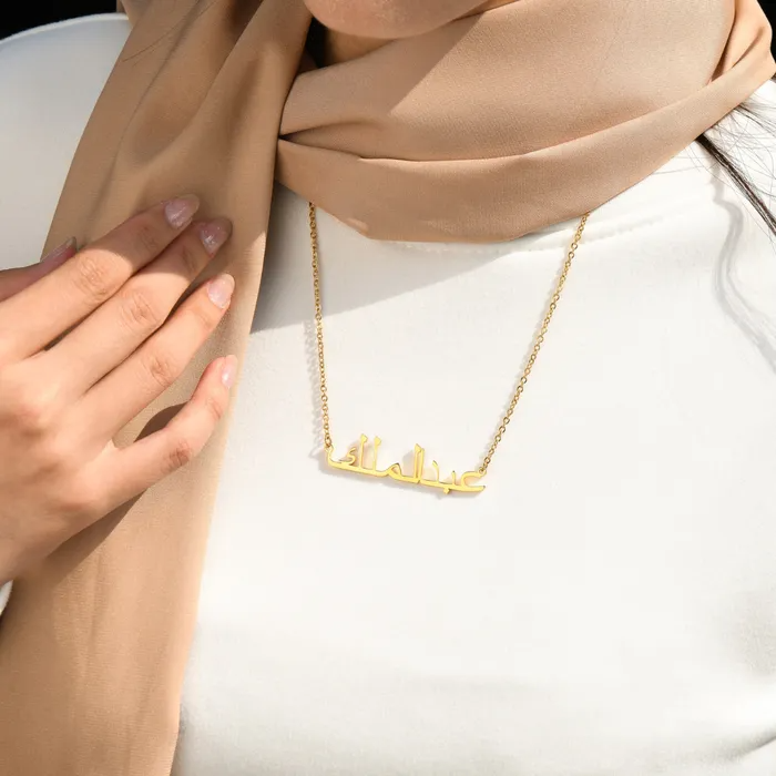 Woman wearing a brown scarf and plain white and a gold Arabic engraved necklace.