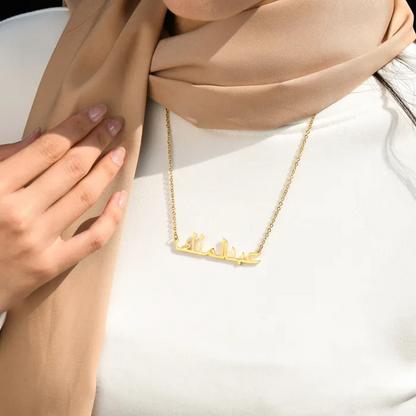 Woman wearing a brown scarf and plain white and a gold Arabic engraved necklace.