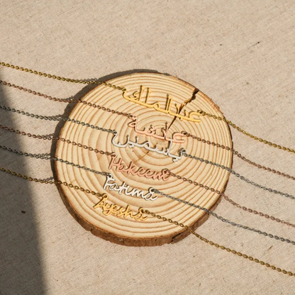 Arabic and English engraved names on gold, rose gold, and silver necklaces on a wooden disc on a plain canvas.