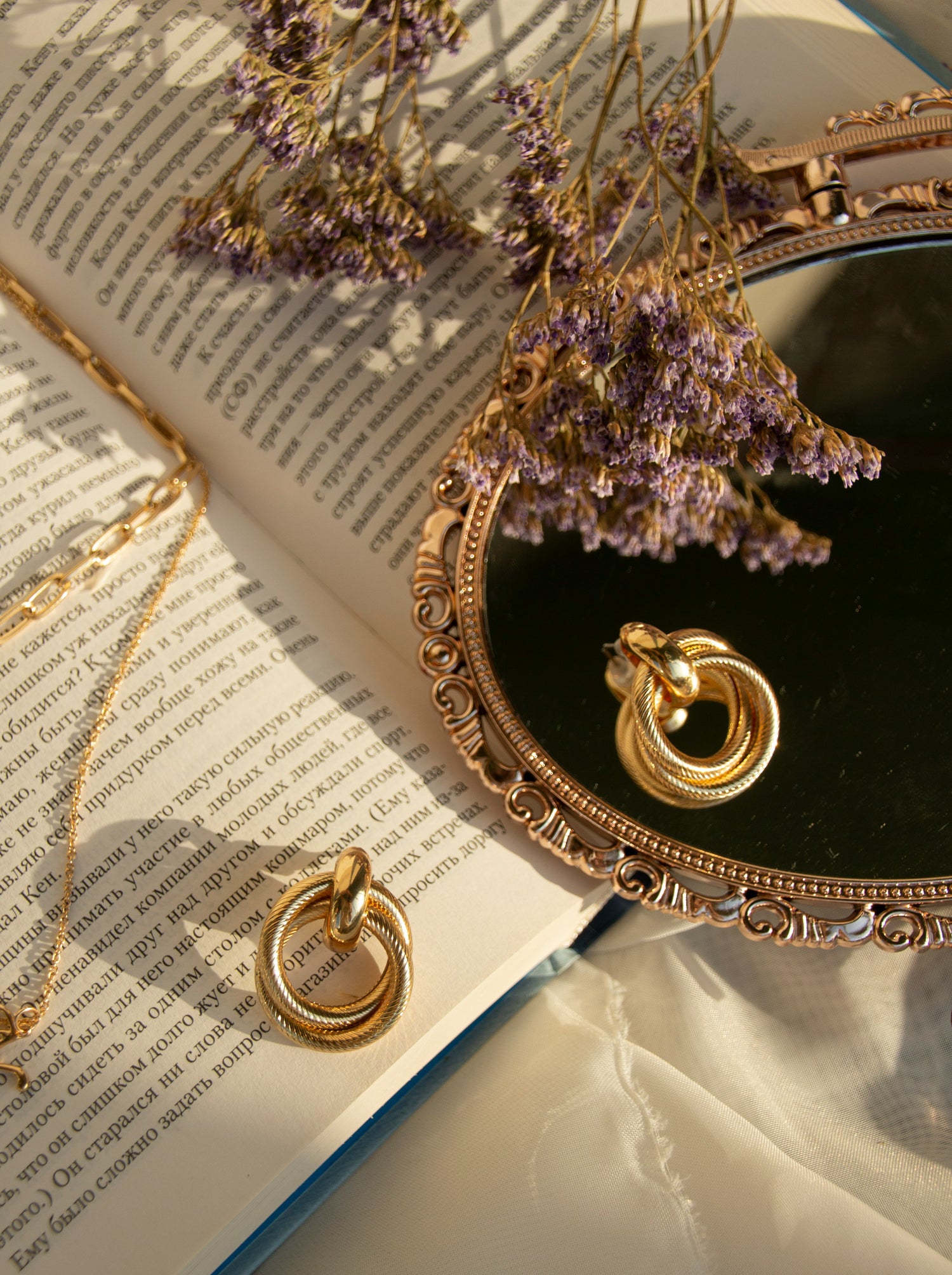 a pair of gold earrings and gold chain necklace, a gold mirror, an open book and delicate purple flowers