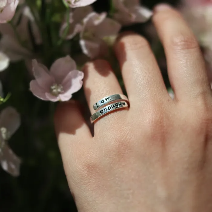 Silver ring with text engraving of "I am enough" on a woman's ring finger with floral background