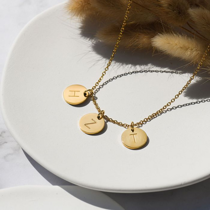 A gold necklace with three gold personalized initial discs on a white circular disc and floral decoration