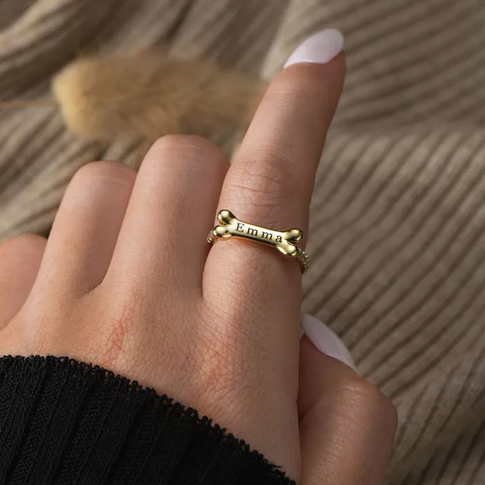 A gold dog bone ring with a personalized name "Emma" on woman's index finger with a knitted beige cloth background