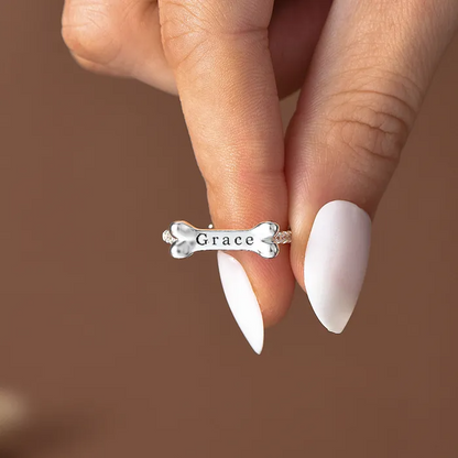 A silver dog bone ring with a personalized name "Grace" held by a woman's fingers with a brown background