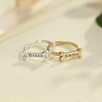 Silver dog bone ring with personalized name "Grace" and gold dog bone ring with personalized name "Emma" on a light background.