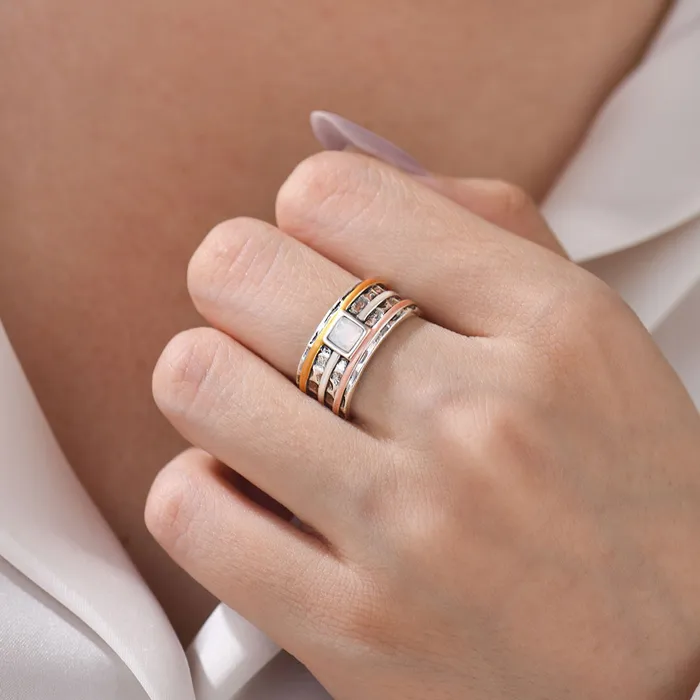 A silver ring with rose gold, silver, and orange bands surrounding it and a pearl-like square stone on woman's middle finger