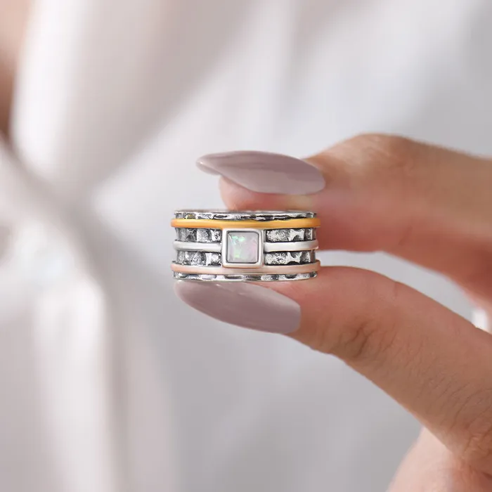 A silver ring with rose gold, silver, and orange bands surrounding it and a pearl-like square stone on woman's fingers