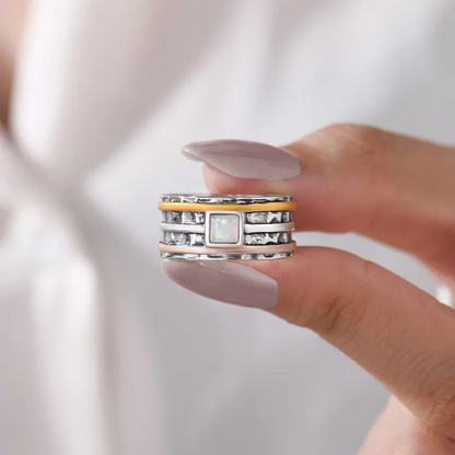 A silver ring with rose gold, silver, and orange bands surrounding it and a pearl-like square stone on woman's fingers