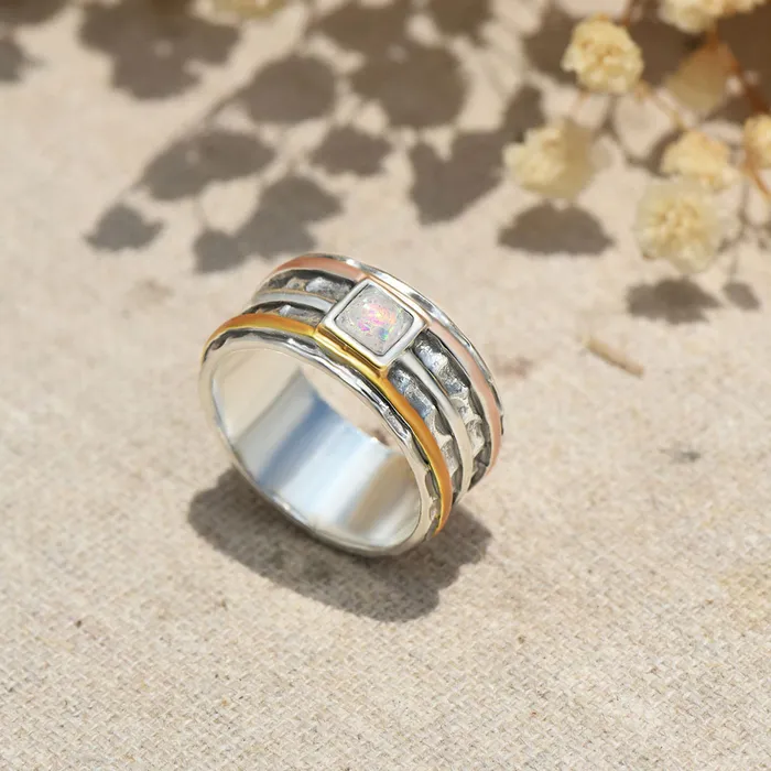 A silver ring with rose gold, silver, and orange bands surrounding it and a pearl-like square stone on top with a floral background