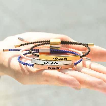 Black rope with gold bar, pink rope with silver bar, and blue rope with rose gold bar each bar with "unbreakable" text engraving stack together on a palm