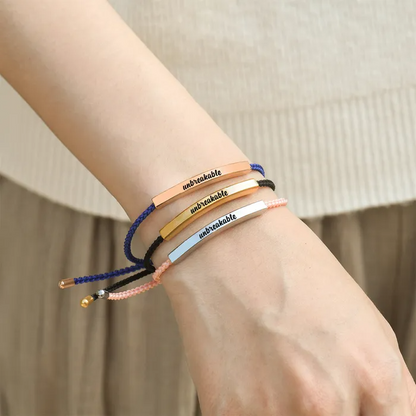 Blue rope with rose gold bar, black rope with gold bar, and pink rope with silver bar each bar has "unbreakable" text engraving on a woman's wrist