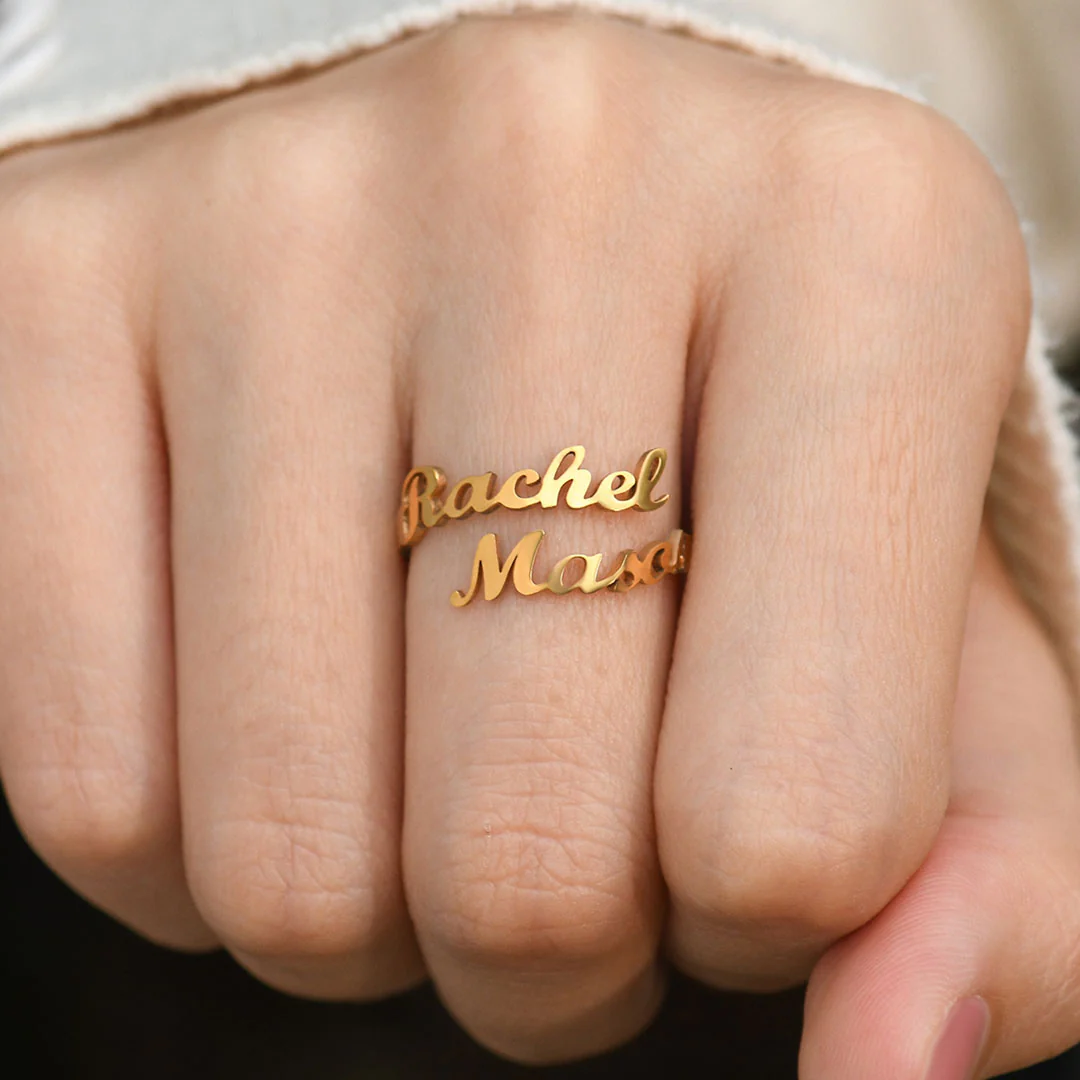 A gold ring featuring two names, "Rachel" and "Mason," elegantly written in a cursive font. The names wrap around the finger, forming the band of the ring in a personalized, stylish design.