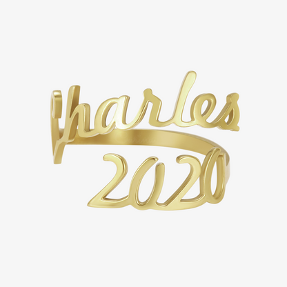 A gold ring featuring two names, "Charles" and "2020," elegantly written in a cursive font. The names wrap around the finger, forming the band of the ring in a personalized, stylish design.