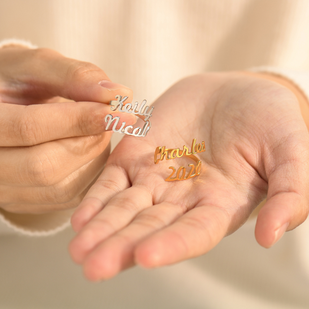 Someone holding a gold and silver ring featuring different names elegantly written in a cursive font. The names wrap around the finger, forming the band of the ring in a personalized, stylish design.