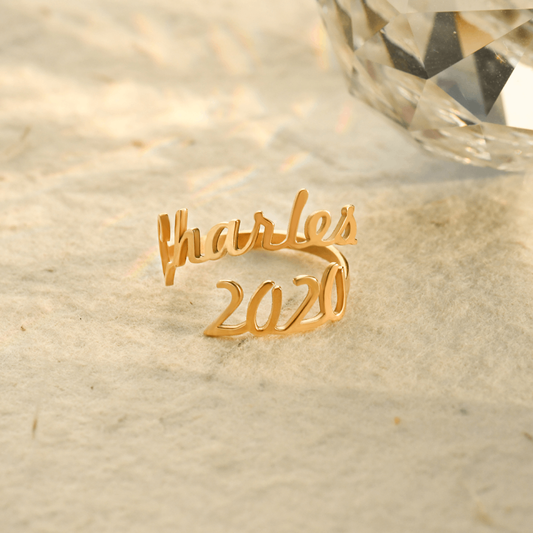 A gold ring featuring two names, "Charles" and "2020," elegantly written in a cursive font. The names wrap around the finger, forming the band of the ring in a personalized, stylish design. The ring is laying on sand 