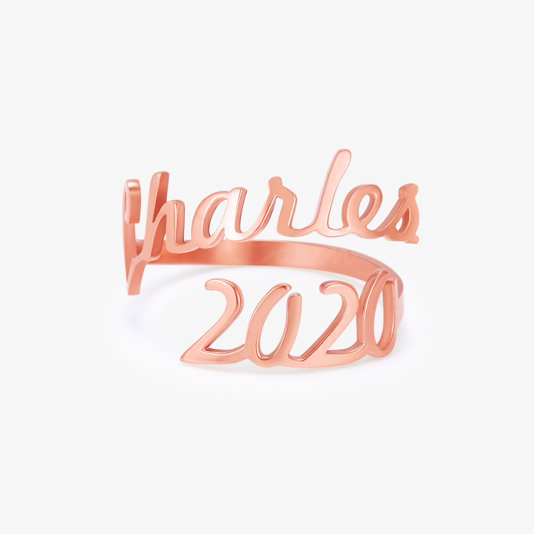 A rose gold ring featuring two names, "Charles" and "2020," elegantly written in a cursive font. The names wrap around the finger, forming the band of the ring in a personalized, stylish design.